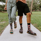 A close paired-feet shot on a brick-edged sidewalk highlights the men's black Pretty Fly For A Fungi socks with red mushrooms beside the women's teal-cuffed pair, both worn with brown buckle sandals. [lf]