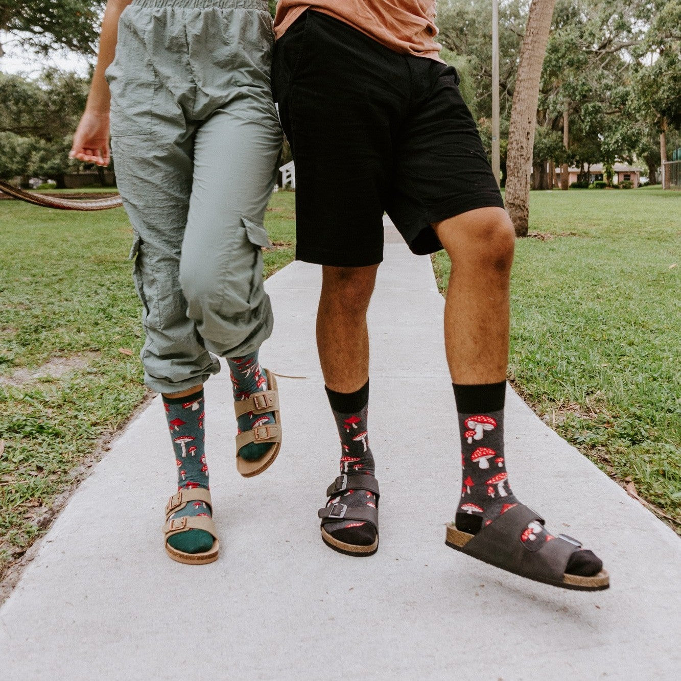 A close paired-feet shot on a brick-edged sidewalk highlights the men's black Pretty Fly For A Fungi socks with red mushrooms beside the women's teal-cuffed pair, both worn with brown buckle sandals. [lf]