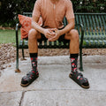 A young man laughs on a green park bench in front of lush palms wearing black Pretty Fly For A Fungi socks with red mushroom prints, paired with a salmon tee, charcoal shorts, and black buckle sandals. [lf]