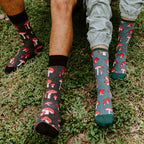 A close shot of two pairs of feet on grass highlights Pretty Fly For A Fungi socks, the women's version with a teal heel and toe beside the men's all-black cuff, both printed with bright red toadstools. [lf]
