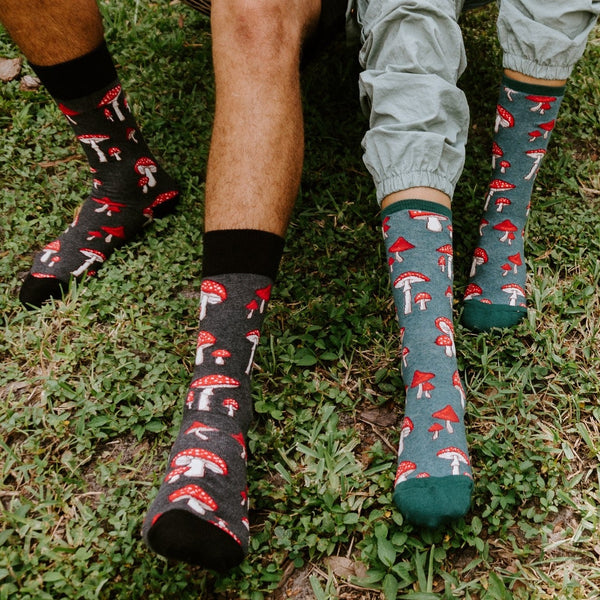 A close shot of two pairs of feet on grass highlights Pretty Fly For A Fungi socks, the women's version with a teal heel and toe beside the men's all-black cuff, both printed with bright red toadstools. [lf]