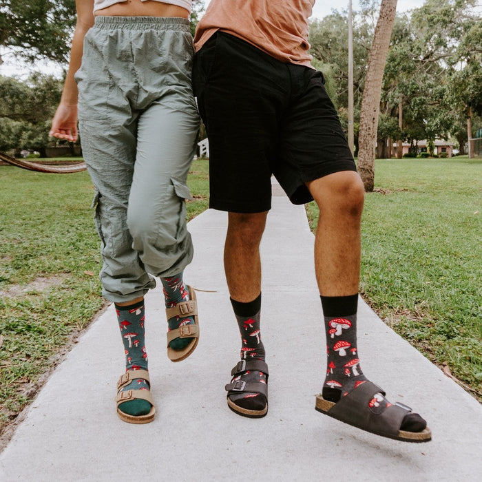 A man and woman laugh together mid-stride on a concrete path, their Pretty Fly For A Fungi socks in Green Heather paired with buckled sandals and casual shorts. [lf]