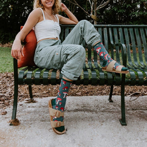A grinning woman sits on a green park bench beside an orange pillow, her Pretty Fly For A Fungi socks in Green Heather peeking above strappy sandals. [lf]