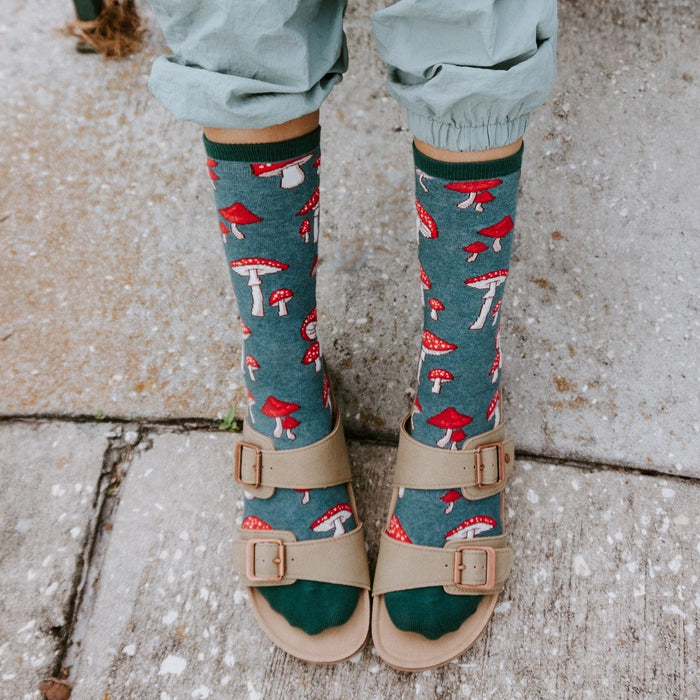 An overhead view of cuffed sage cargo pants over Pretty Fly For A Fungi socks in Green Heather scattered with red toadstools, tucked into tan two-strap buckle sandals on a stone patio. [lf]