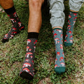 Two models stand on a grassy patch in matching Pretty Fly For A Fungi socks in Green Heather, the mushroom print visible at calf height above their ankles. [lf]
