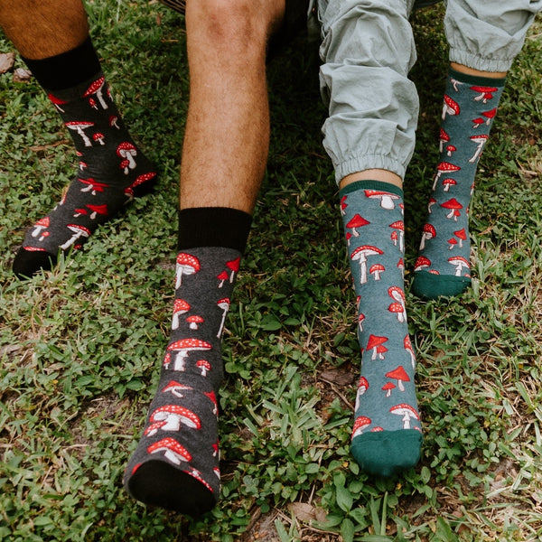 Two models stand on a grassy patch in matching Pretty Fly For A Fungi socks in Green Heather, the mushroom print visible at calf height above their ankles. [lf]