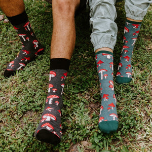 Two models stand on a grassy patch in matching Pretty Fly For A Fungi socks in Green Heather, the mushroom print visible at calf height above their ankles. [lf]