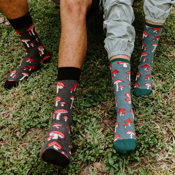 Two models stand on a grassy patch in matching Pretty Fly For A Fungi socks in Green Heather, the mushroom print visible at calf height above their ankles. [lf]