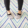 An overhead view of feet standing on a blue and teal chevron rug, The Cats Meow no-show socks peeking out with gray cat faces above black leggings. [lf]