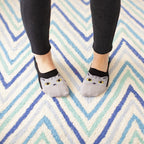 An overhead view of feet standing on a blue and teal chevron rug, The Cats Meow no-show socks peeking out with gray cat faces above black leggings. [lf]
