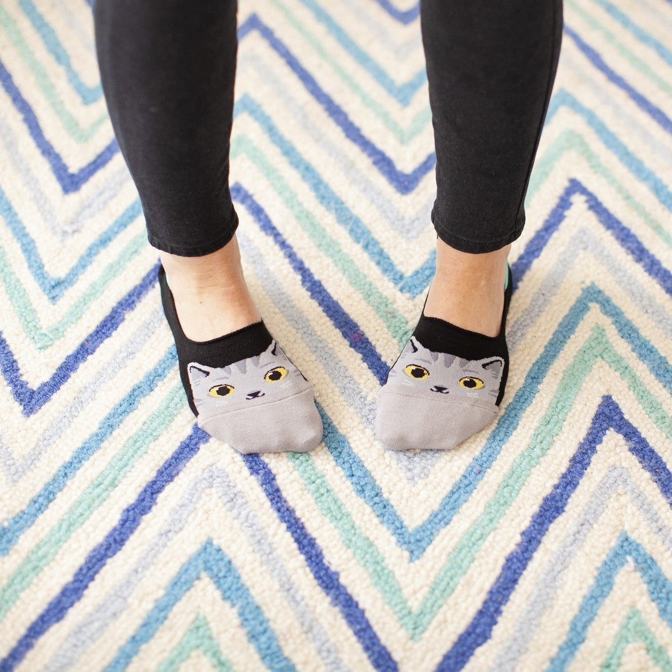 An overhead view of feet standing on a blue and teal chevron rug, The Cats Meow no-show socks peeking out with gray cat faces above black leggings. [lf]