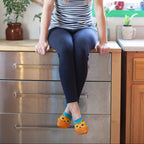 A bright kitchen scene with a model perched on a steel counter in stripes and leggings, swinging legs that show off The Cats Meow socks with orange tabby cat faces. [lf]