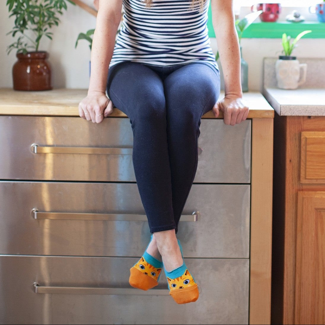 A bright kitchen scene with a model perched on a steel counter in stripes and leggings, swinging legs that show off The Cats Meow socks with orange tabby cat faces. [lf]