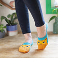 A wide close-up of mid-stride feet on a wooden floor among houseplants, the orange tabby cat faces of The Cats Meow socks tucked low under skinny black pants. [lf]