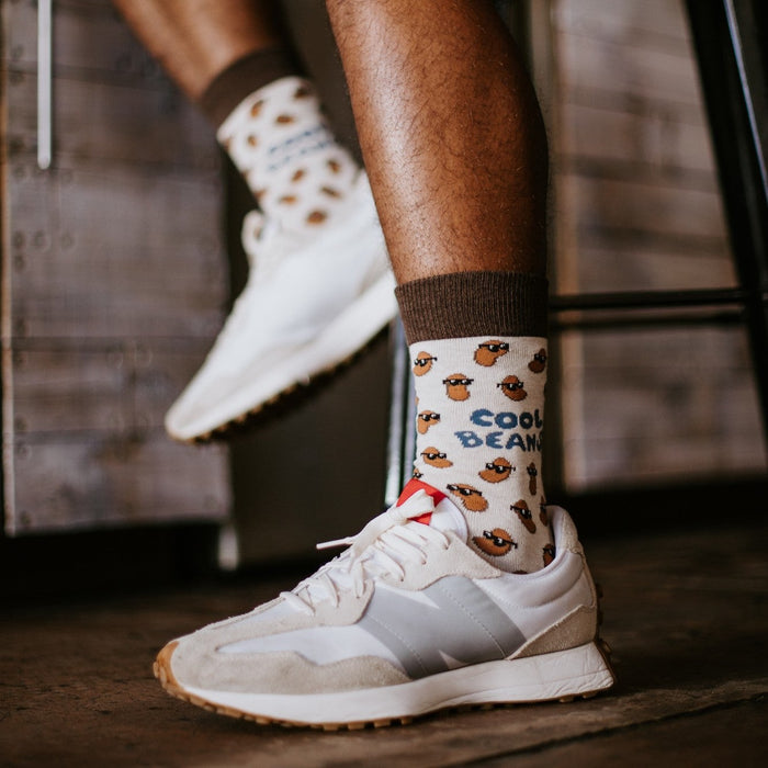 Man perched on a stool in tan shorts swings one leg forward, his planted foot wearing a Cool Beans sock in Ivory Heather tucked into a white sneaker with the blue Cool Beans graphic facing the camera. [lf]
