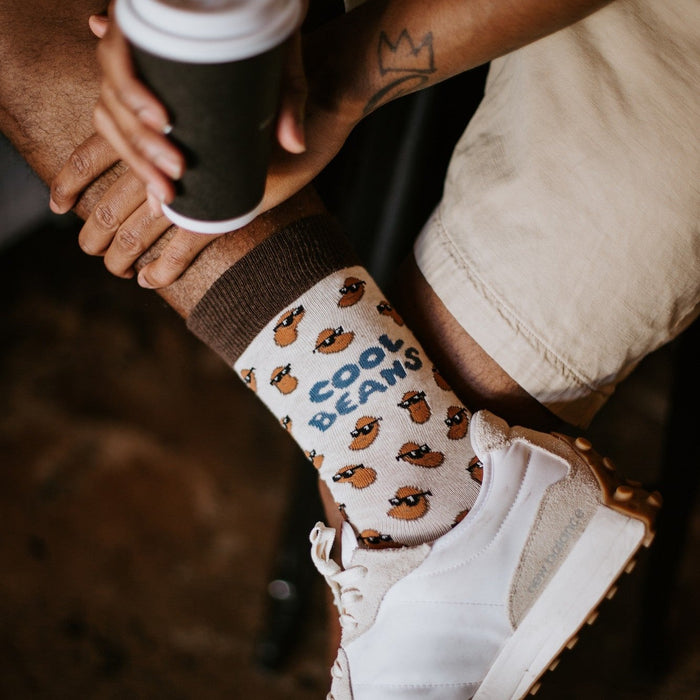 Close detail of a man holding a takeaway coffee cup, crown tattoo on his wrist, ankle resting on his knee to show off a Cool Beans sock in Ivory Heather with brown trim, smiling beans, and blue script lettering above a white sneaker. [lf]