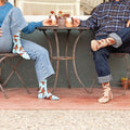 Mid-laugh moment between two friends leaning back in their bistro chairs, mugs hovering over a tiny round table, butterfly-printed Social Butterfly socks visible above his cuffed jeans and her pulled-up denim hem. [lf]