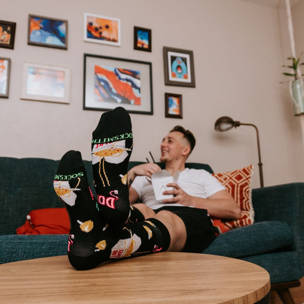 Low-angle view across a round wood table shows crossed feet kicked up on the surface wearing 24 Hour Diner socks in Black, with a relaxed model holding a takeout box behind on the teal couch in a softly lit living room. [lf]