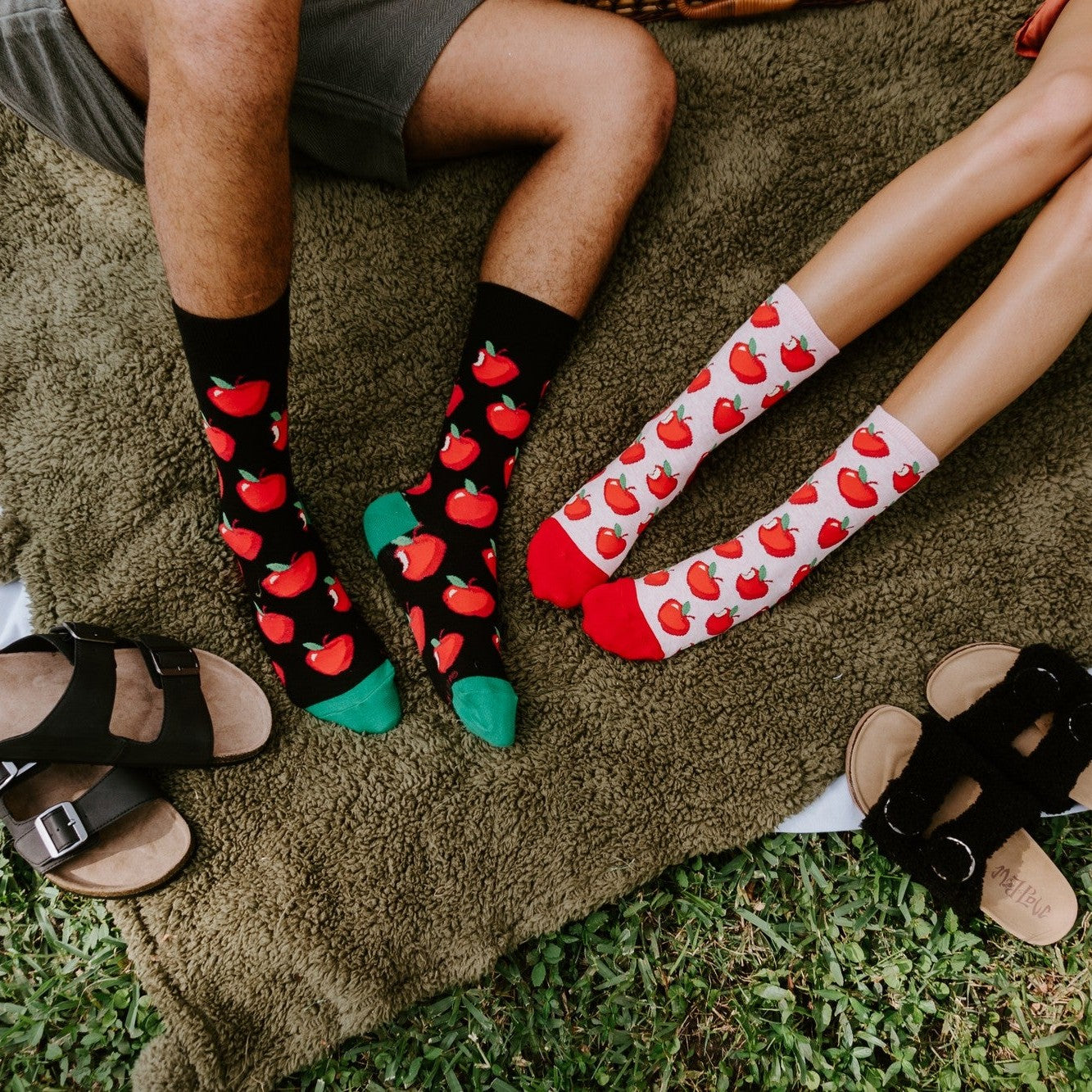 An overhead crop of a couple's feet on a green picnic rug with sandals kicked off beside them, the man wearing the black Apple Of My Eye socks with green toes and the woman wearing the white Apple Of My Eye socks with red apples and red soles. [lf]