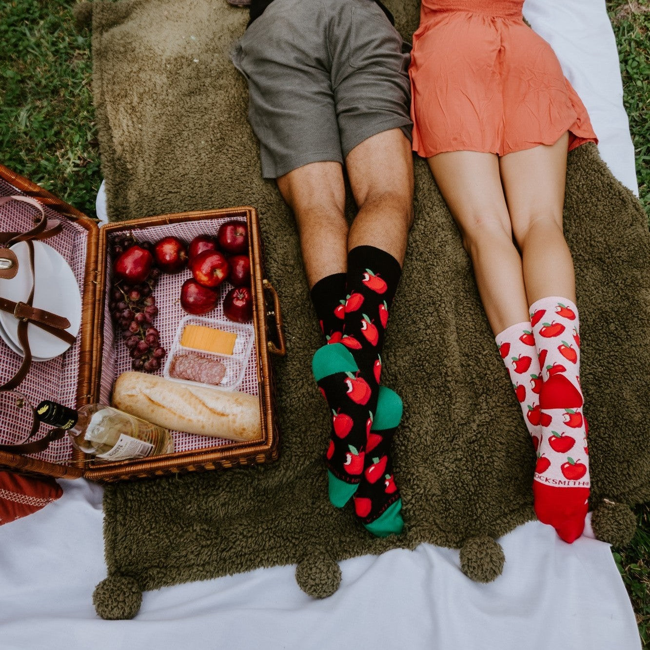 An overhead view of a couple lying side by side on a green picnic blanket beside an open wicker basket of apples, cheese and bread; he wears the black Apple Of My Eye socks and she wears the white Apple Of My Eye socks, both patterned with red apples. [lf]