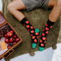 A man in grey shorts sits cross-legged on a fluffy green picnic blanket beside an open wicker basket of red apples, salami and cheese, wearing the black Apple Of My Eye socks with red apple prints and bright green toes. [lf]
