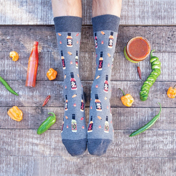 Flat-lay overhead of feet on weathered wood planks surrounded by scattered habaneros, jalapenos and a hot sauce bottle, wearing the Hot In Here socks in slate blue printed with tiny hot sauce bottles and chili peppers. [lf]