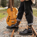 Standing on a railroad track in cuffed black jeans and chunky leather boots, a person in a plaid flannel balances a vintage acoustic guitar by the neck beside their leg in Nice Acoustics Bamboo socks in Charcoal Heather. [lf]