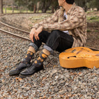 Profile of a guitarist sitting trackside under bare winter branches, knees drawn up and acoustic guitar leaned against one leg, with rolled denim revealing Nice Acoustics Bamboo socks in Charcoal Heather. [lf]