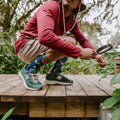 A man in a bucket hat crouches and peers through a magnifying glass into the foliage on a sunlit deck while wearing Brilliant Bugs Bamboo socks in Navy Heather with green New Balance sneakers. [lf]