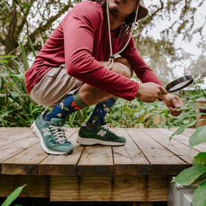 A man in a bucket hat crouches and peers through a magnifying glass into the foliage on a sunlit deck while wearing Brilliant Bugs Bamboo socks in Navy Heather with green New Balance sneakers. [lf]
