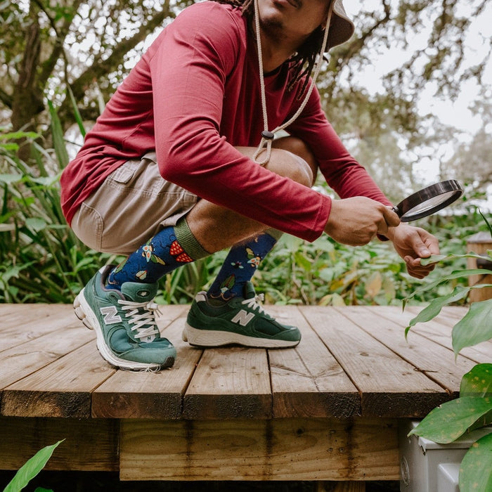 A man in a bucket hat crouches and peers through a magnifying glass into the foliage on a sunlit deck while wearing Brilliant Bugs Bamboo socks in Navy Heather with green New Balance sneakers. [lf]