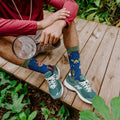 A close cropped shot framing only the legs and torso of a seated man holding a magnifying glass, the Brilliant Bugs Bamboo socks in Navy Heather contrasting brightly against green New Balance sneakers on a wood deck. [lf]