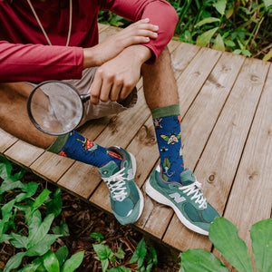 A close cropped shot framing only the legs and torso of a seated man holding a magnifying glass, the Brilliant Bugs Bamboo socks in Navy Heather contrasting brightly against green New Balance sneakers on a wood deck. [lf]