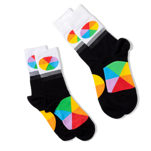 Two pairs of black crew socks with white cuffs and color wheel designs, lying flat and angled differently on a black background.