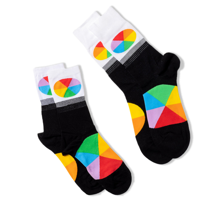 Two pairs of black crew socks with white cuffs and color wheel designs, lying flat and angled differently on a black background.
