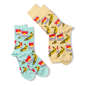 Two pairs of pop art crew socks: one light blue and one light yellow, both featuring red soup cans and yellow bananas, against a black background.