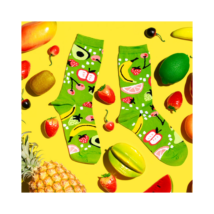 Flat lay of fruit-patterned lime green socks surrounded by fresh and artificial fruits on a vibrant yellow background.
