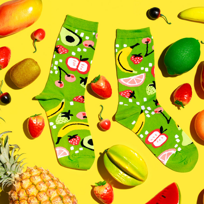 Flat lay of fruit-patterned lime green socks surrounded by fresh and artificial fruits on a vibrant yellow background.