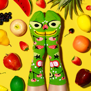 Feet wearing fruit-patterned lime green socks, crossed to make a fruit face, surrounded by scattered fruits on a yellow surface.