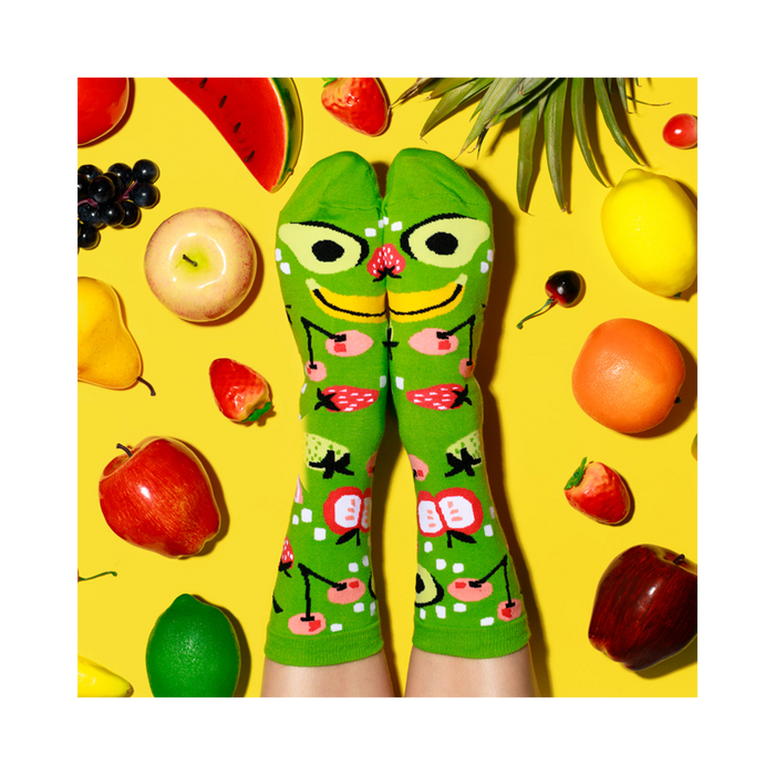 Feet wearing fruit-patterned lime green socks, crossed to make a fruit face, surrounded by scattered fruits on a yellow surface.