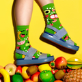 Person's legs wearing fruit-patterned lime green socks and blue sandals, standing on a pile of diverse fruits on yellow. [lf]