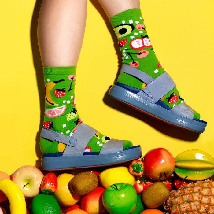 Person's legs wearing fruit-patterned lime green socks and blue sandals, standing on a pile of diverse fruits on yellow.