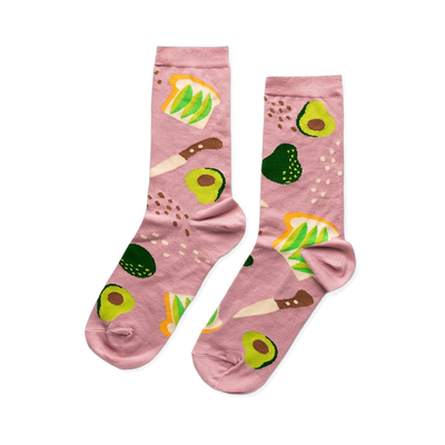 A pair of pink crew socks patterned with avocado toast, sliced and whole avocados, and a knife, on a light pink background.