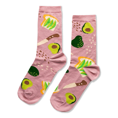 A pair of pink crew socks patterned with avocado toast, sliced and whole avocados, and a knife, on a light pink background.