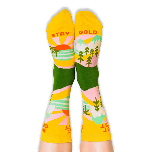 Person wearing the yellow and green socks with their feet up, revealing the words Stay Gold printed on the bottom soles.