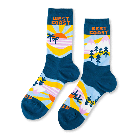 A pair of mismatched crew socks with West Coast beach scenes (sun, palm trees) and Best Coast mountain scenes (peaks, pine trees) on a dark teal base.