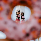 Tight detail of feet in Delicious Donuts Bamboo socks in Black with pink cuffs and white sneakers framed through the hole of a giant pink-frosted donut held close to the camera. [lf]