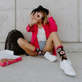 Woman peeks through a pink-frosted donut held to her eye, seated cross-legged in shorts and a hot pink shirt, showing off Delicious Donuts Bamboo socks in Black paired with white sneakers next to an open donut box. [lf]