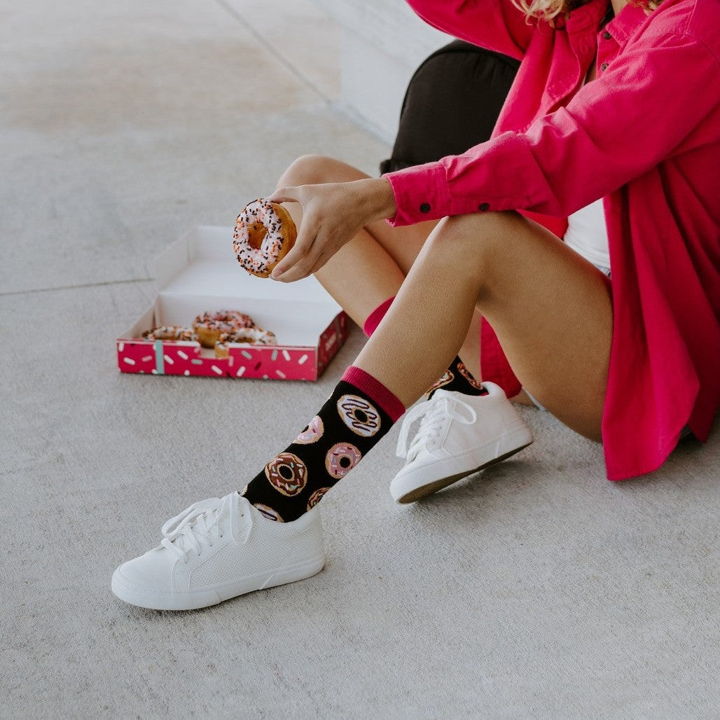 Smiling woman in sunglasses and a wide-brim hat sits against a concrete wall holding a donut, dressed in a hot pink shirt and white shorts with Delicious Donuts Bamboo socks in Black tucked into white sneakers. [lf]