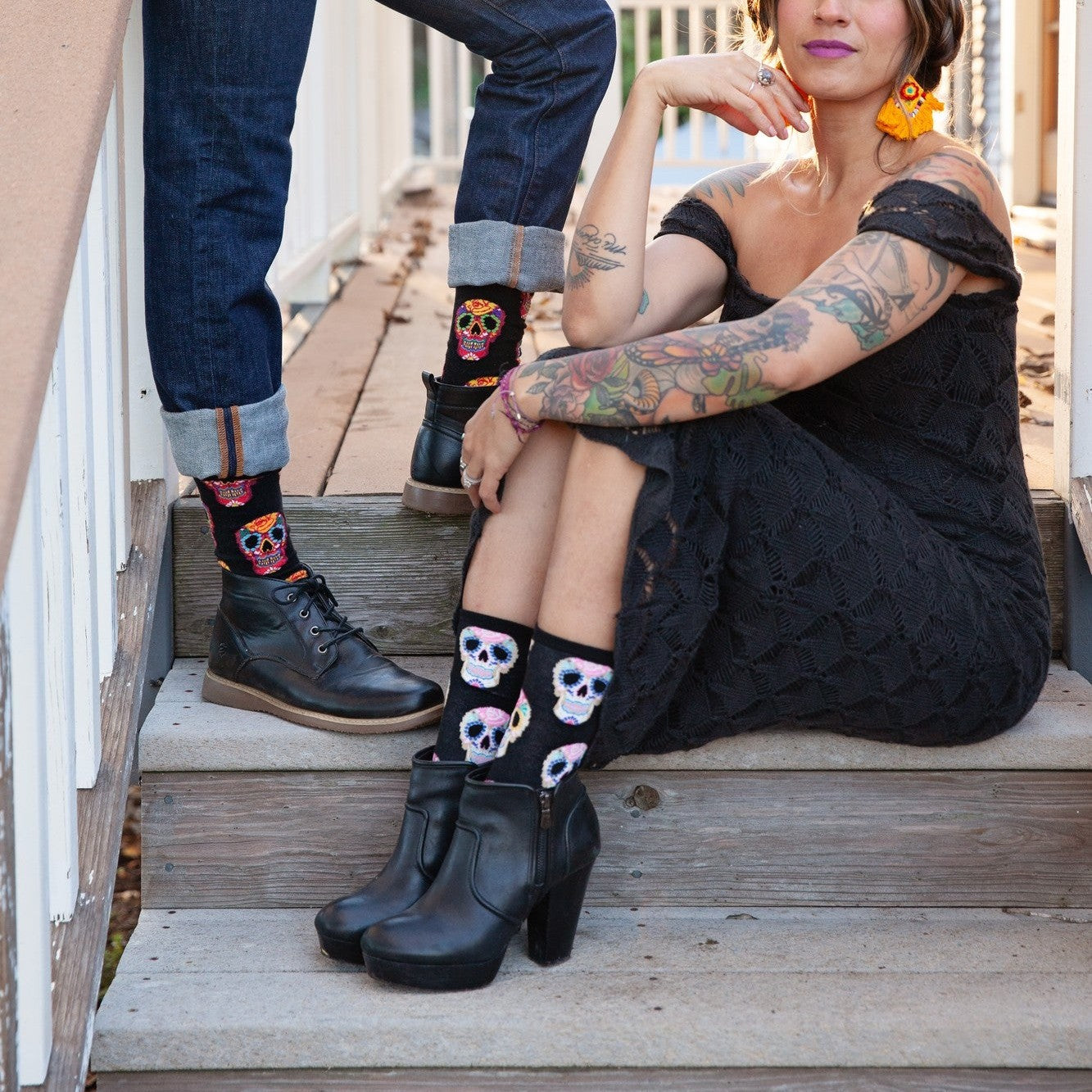 Tattooed woman in a black lace dress sits on a wooden porch step at golden hour beside a man in cuffed jeans and brown leather boots, both wearing Dia De Los Muertos Skull socks in Black with vibrant sugar-skull motifs. [lf]